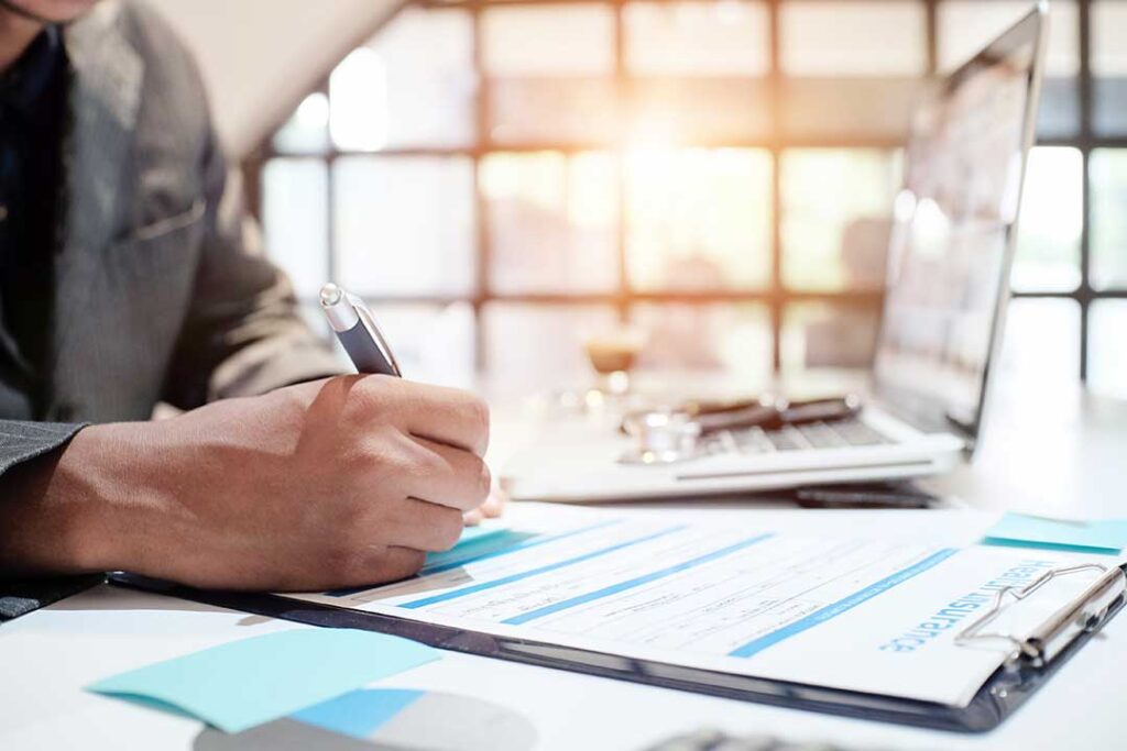 A young adult reviews paperwork at a home kitchen table with a laptop and mug nearby.